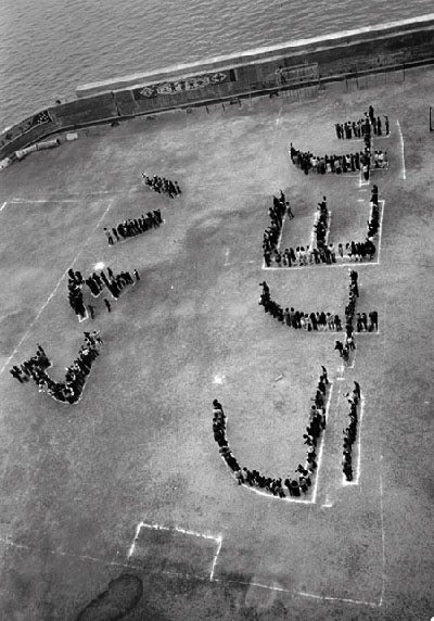 Aerial black-and-white photo of crowd forming “SAVE US” beside a waterfront seawall.