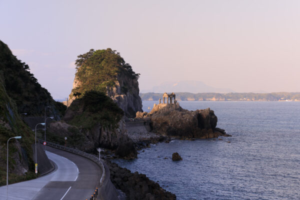 Tranquil coastal beauty with winding road, rock formations, lush foliage, and striking temple.