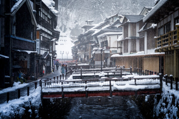 Winter charm of Ginzan Onsen: traditional buildings, snow-covered canal, cozy ambiance, falling snowflakes.