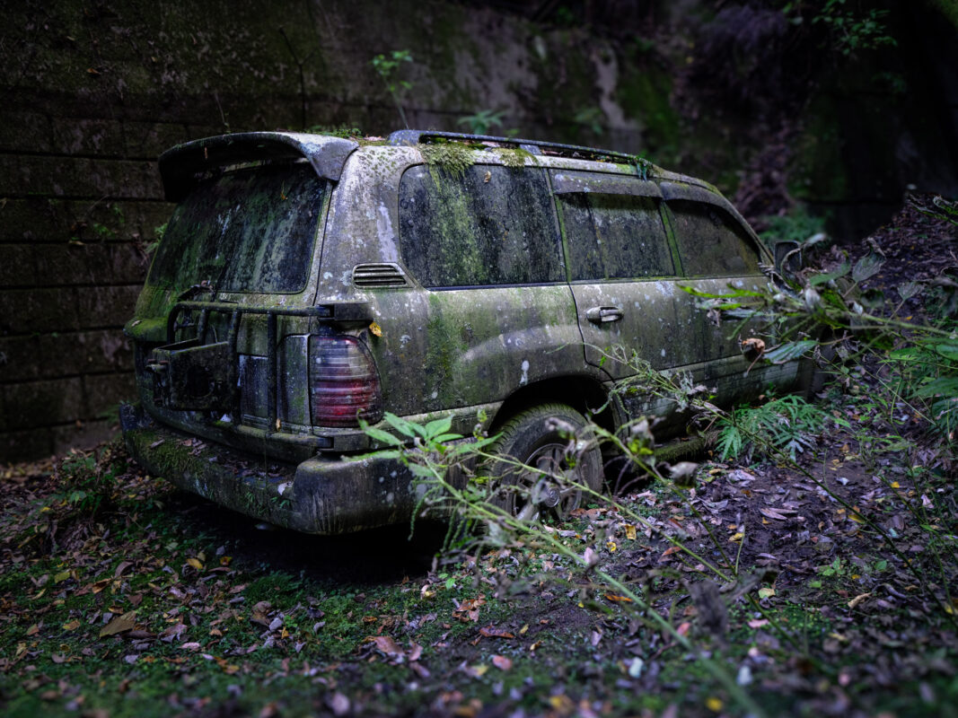 Abandoned Toyota Land Cruiser covered in moss in a forest ravine near a retaining wall.