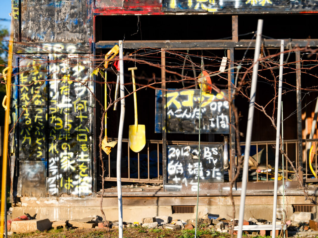 Abandoned black house facade with graffiti, rusted barred windows, and tangled wires in sunlight.