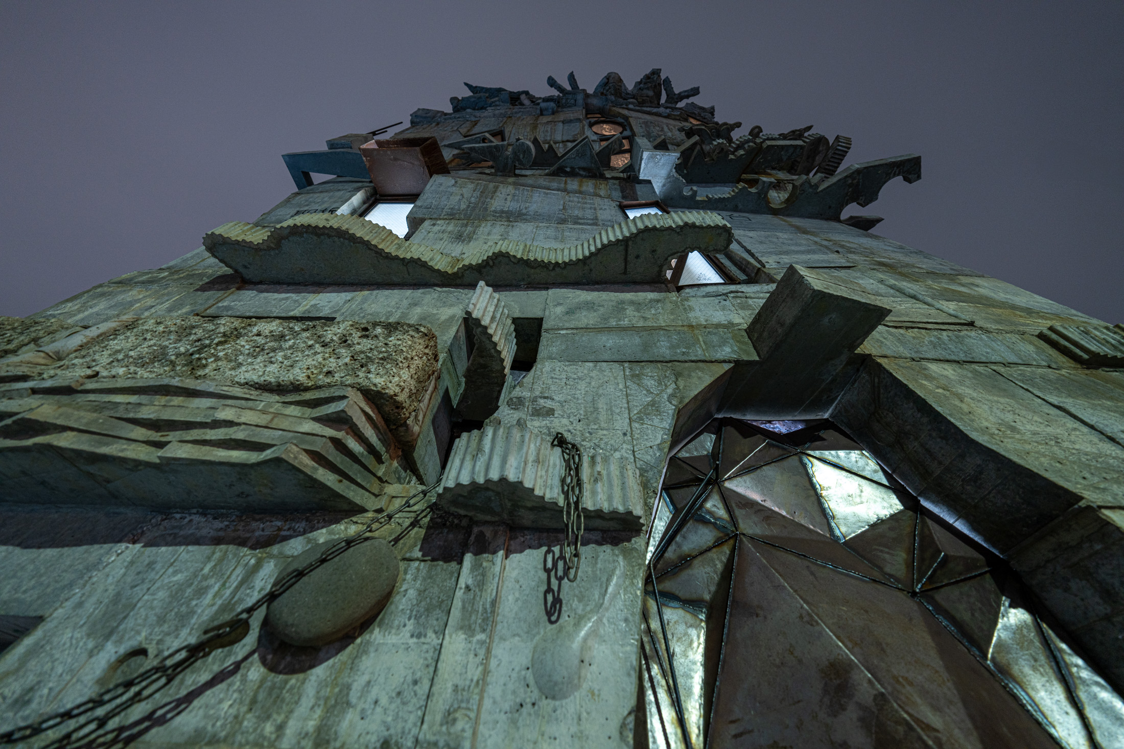 Low-angle brutalist megastructure tower with weathered metal and stone against gray-blue sky