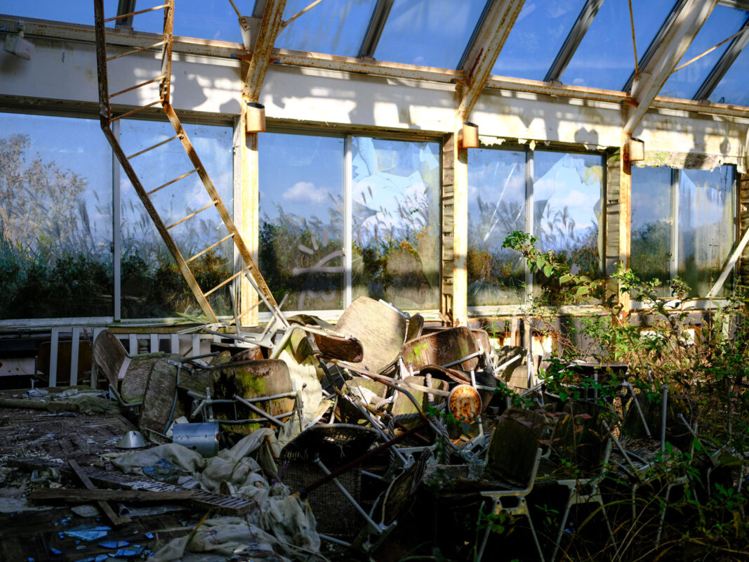 Sunlit abandoned glass church ruins with broken chairs, debris, and plants reclaiming the interior.