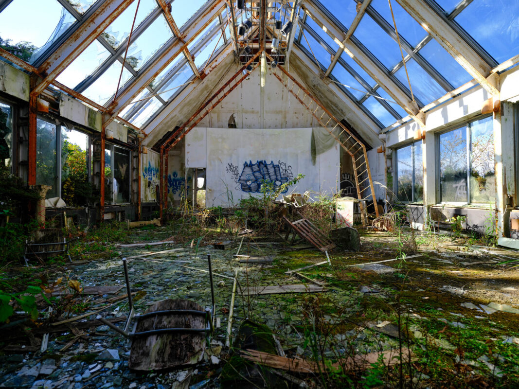 Abandoned glass-roof chapel ruins with graffiti, broken panes, and weeds in sunlight.