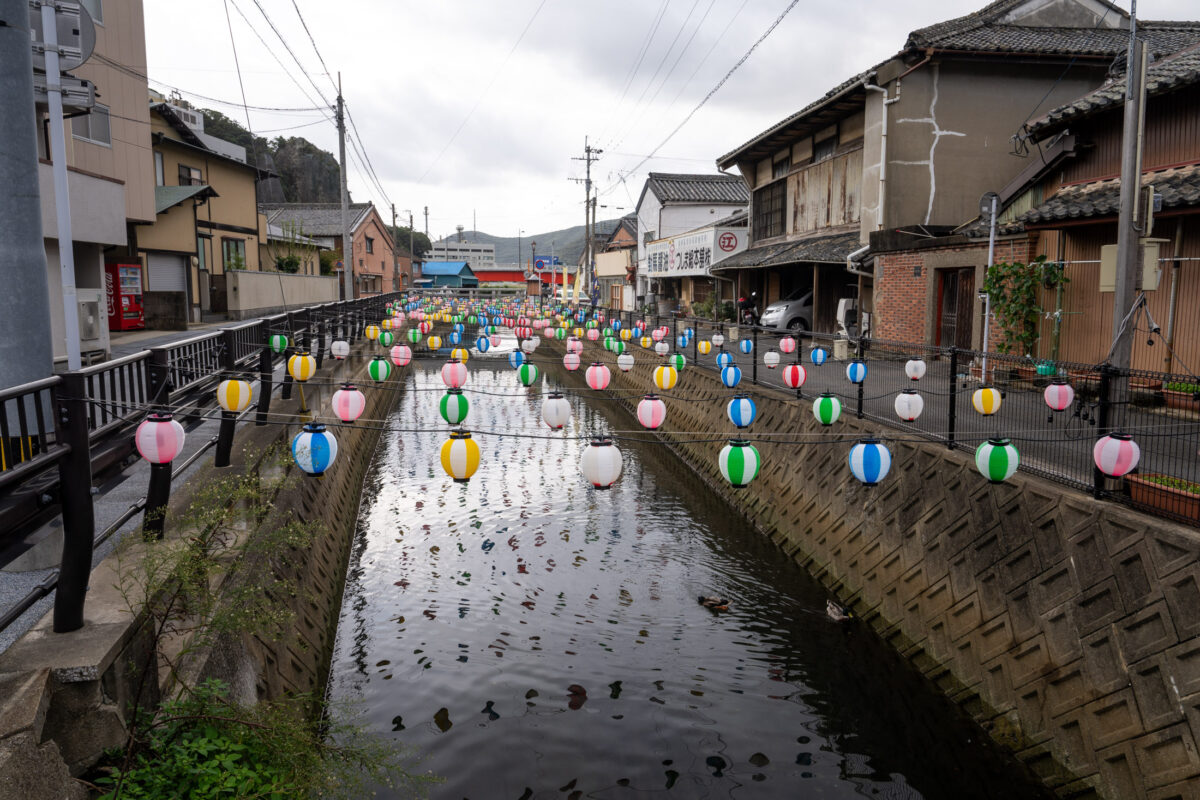Pastel lanterns over Izuhara canal in Japan, reflecting on rippling water under cloudy skies.