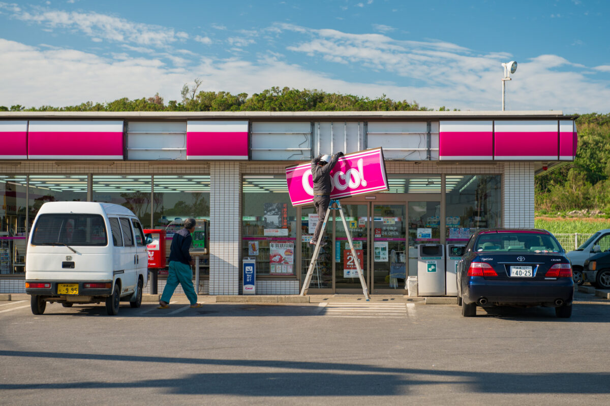 Workers on ladder repairing pink coco! sign at Miyako Island roadside shop, Japan.