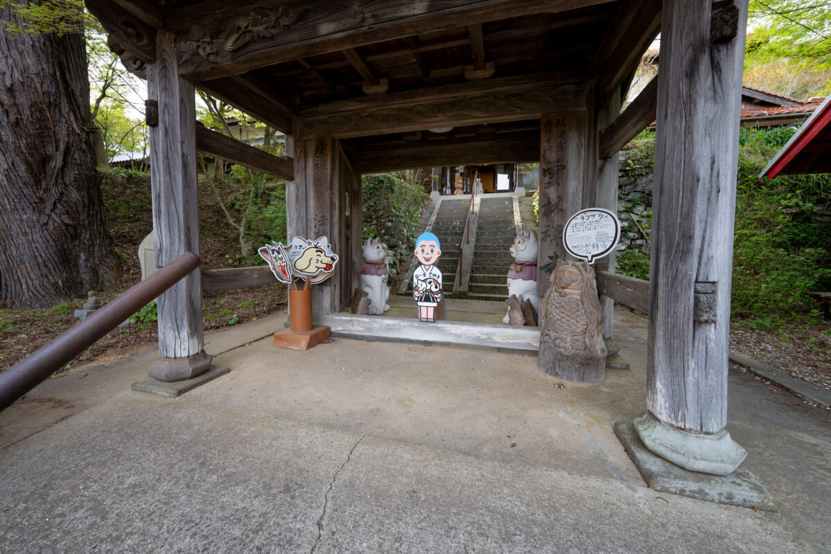 Unrin-ji temple gate in Japan with guardian statues, stone steps, and cartoon monk cutout.