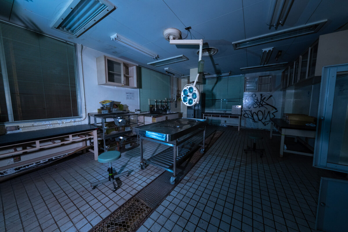 Abandoned animal research lab with surgical table, stainless steel carts, and graffiti under fluorescent lights.