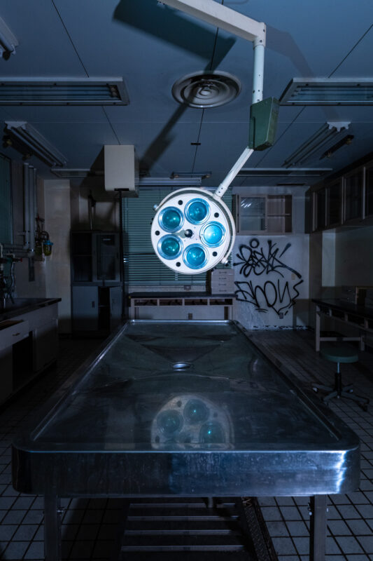 Abandoned animal research lab with stainless surgical table and blue examination lamp, graffiti visible.