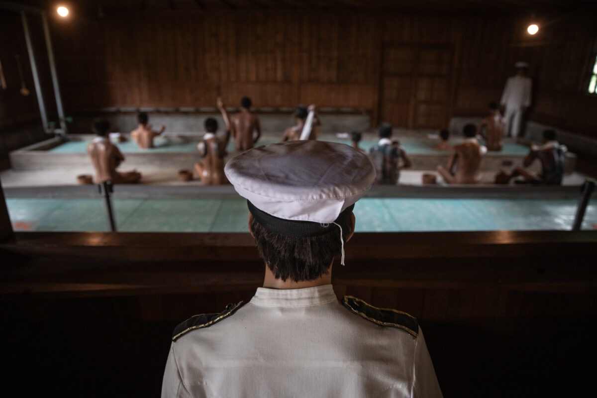 Abashiri Prison bathing hall interior, guard overlooking inmates soaking in communal bath.