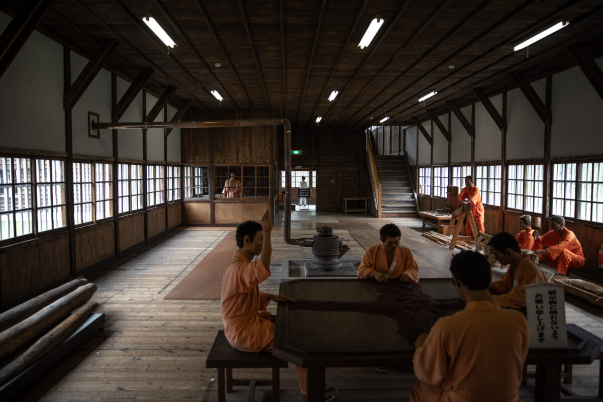 Abashiri Prison interior hall with inmates in orange uniforms working at wooden tables
