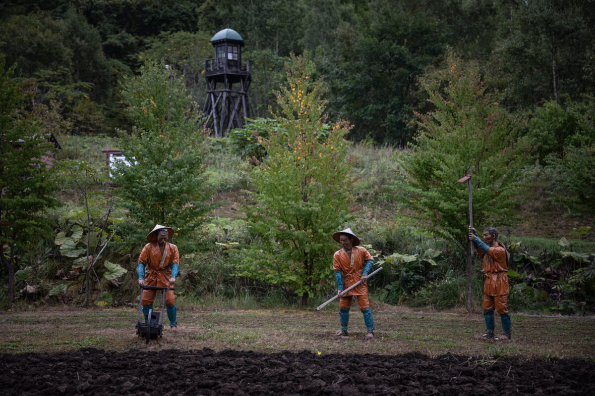 Abashiri Prison laborers till field with hand tools under watchtower in rural Japan.