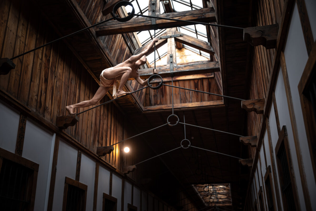 Suspended nude figure in Abashiri Prison wooden corridor with cables and skylight shadows.