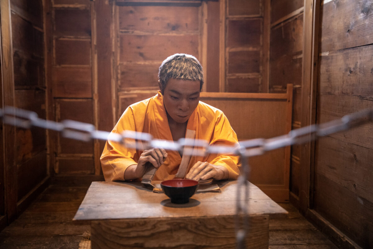 Inmate eating simple meal inside Abashiri Prison wooden cell, chain barrier in foreground.