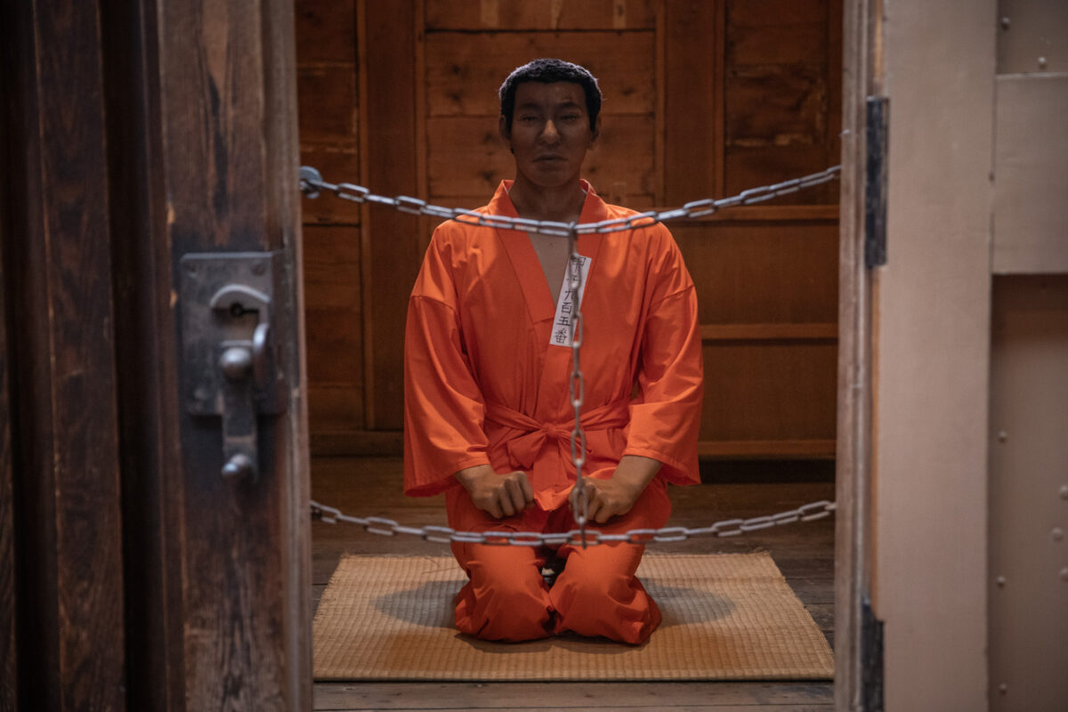 Abashiri Prison wooden cell with chained doorway and kneeling prisoner in orange uniform