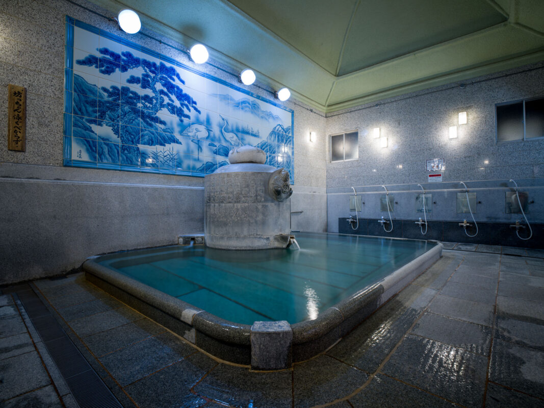 Interior of Dōgo Onsen bathhouse with steaming stone bath, blue mural, and washing stations
