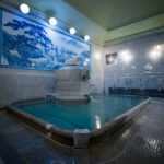 Interior of Dōgo Onsen bathhouse with steaming stone bath, blue mural, and washing stations
