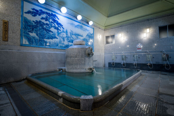 Interior of Dōgo Onsen bathhouse with steaming stone bath, blue mural, and washing stations