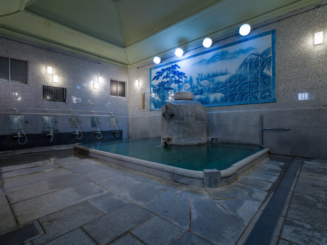 Dogo Onsen bathing hall with stone hot spring pool, washing stations, and scenic mural