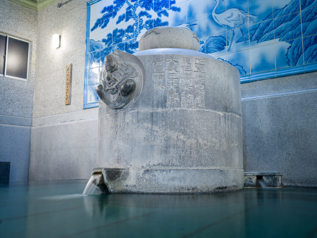 Dogo Onsen indoor hot spring bath with carved stone pillar fountain and blue tile mural.
