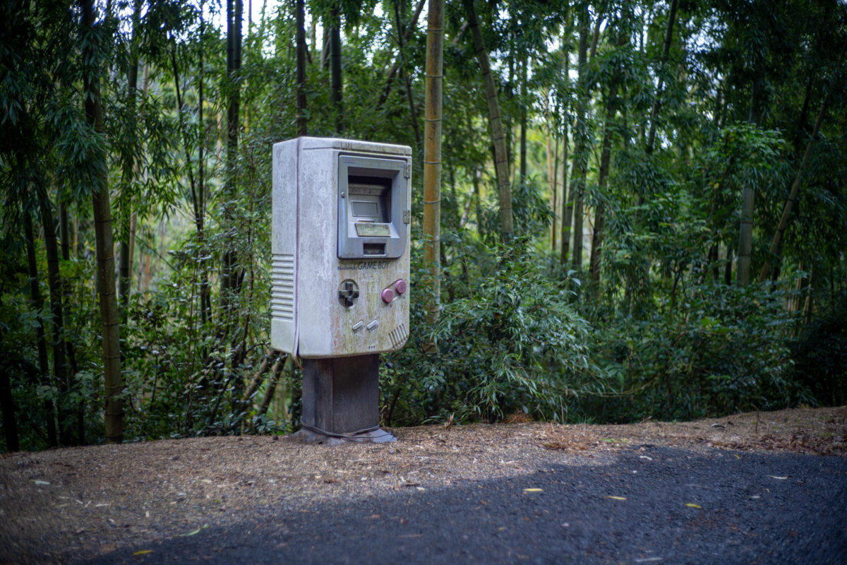 Giant weathered Game Boy mailbox beside a paved forest path among dense green trees.