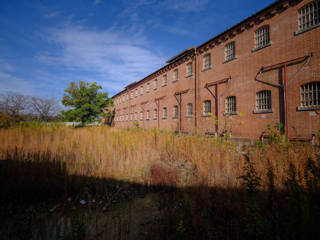 Abandoned Nara Prison brick wall with barred windows and overgrown grass under blue sky.