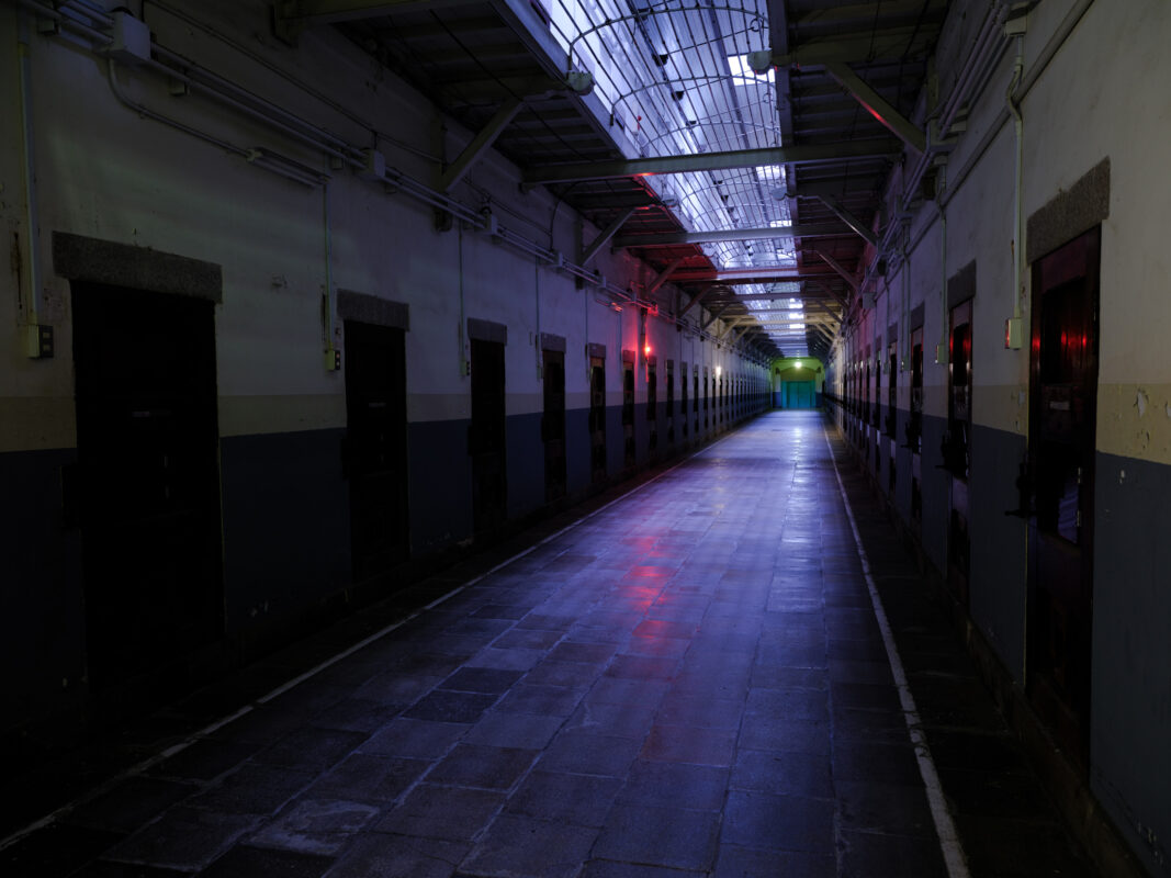 Empty Nara Prison corridor with symmetrical cell doors, cold overhead light, and red warning lights