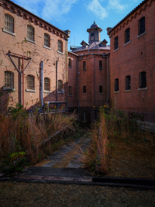 Abandoned Nara Prison courtyard with red-brick walls, barred windows, and overgrown path