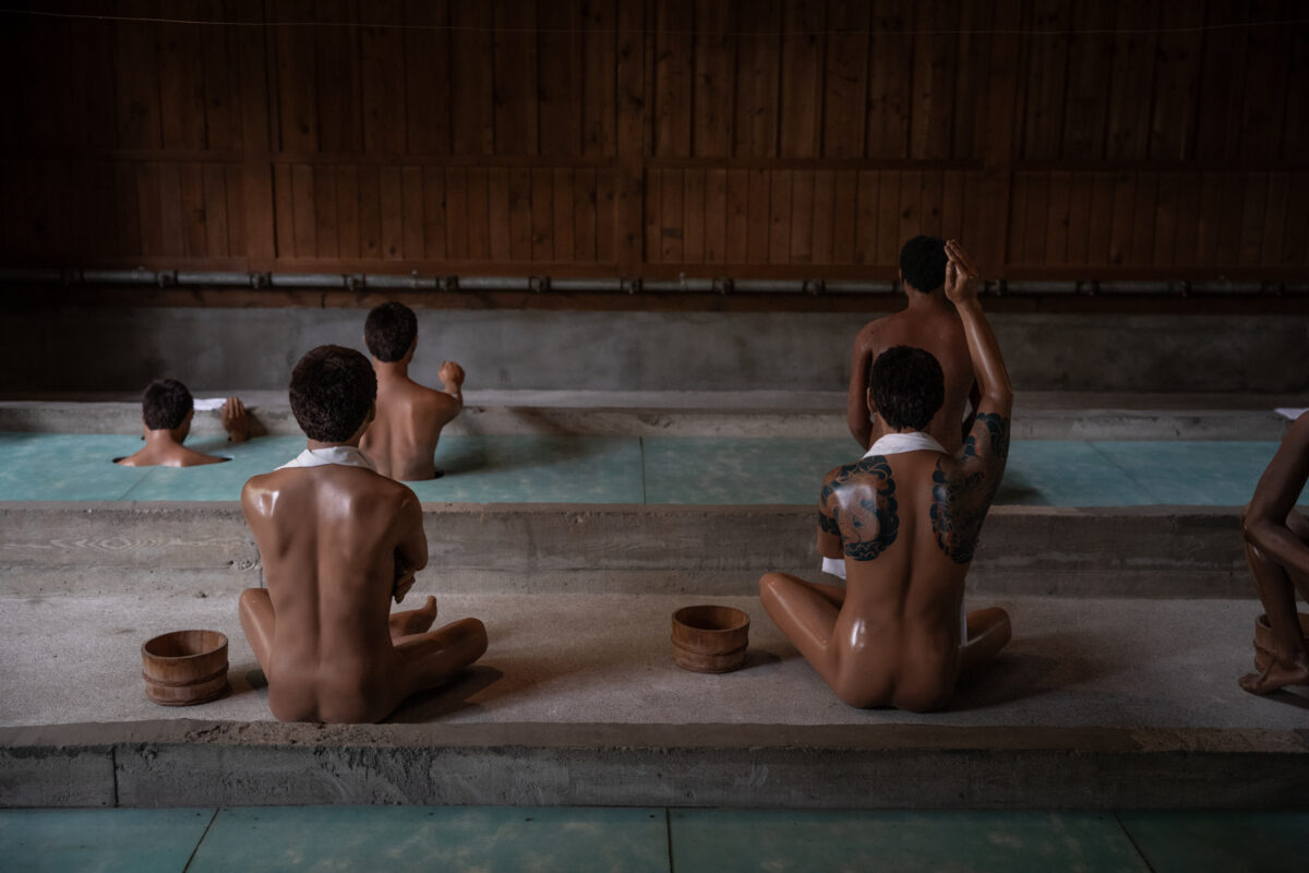 Inmates seated in Abashiri Prison communal bathhouse beside indoor pool and wooden buckets.